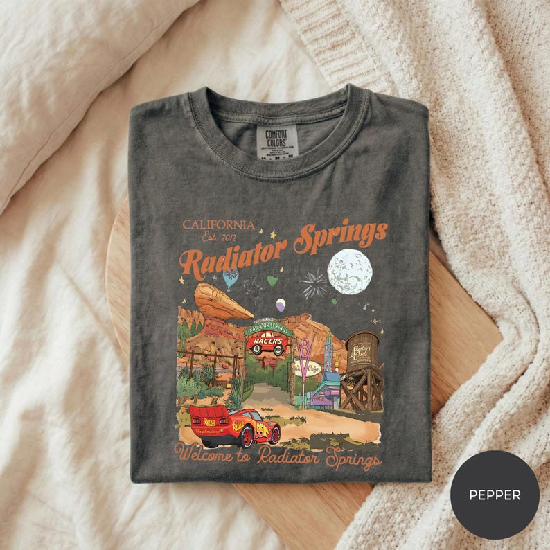 A t-shirt with a graphic design depicting a desert landscape with a red car, a sign that says "RADIATOR SPRINGS", and various other elements associated with the location.