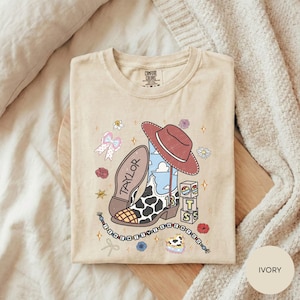 A beige t-shirt with a cartoon-style illustration of a cowboy boot, a red hat, and various other cartoon-style elements, along with the word "TAYLOR" printed on it.