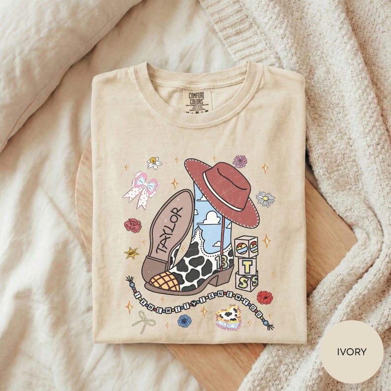 A beige t-shirt with a cartoon-style illustration of a cowboy boot, a red hat, and various other cartoon-style elements, along with the word "TAYLOR" printed on it.