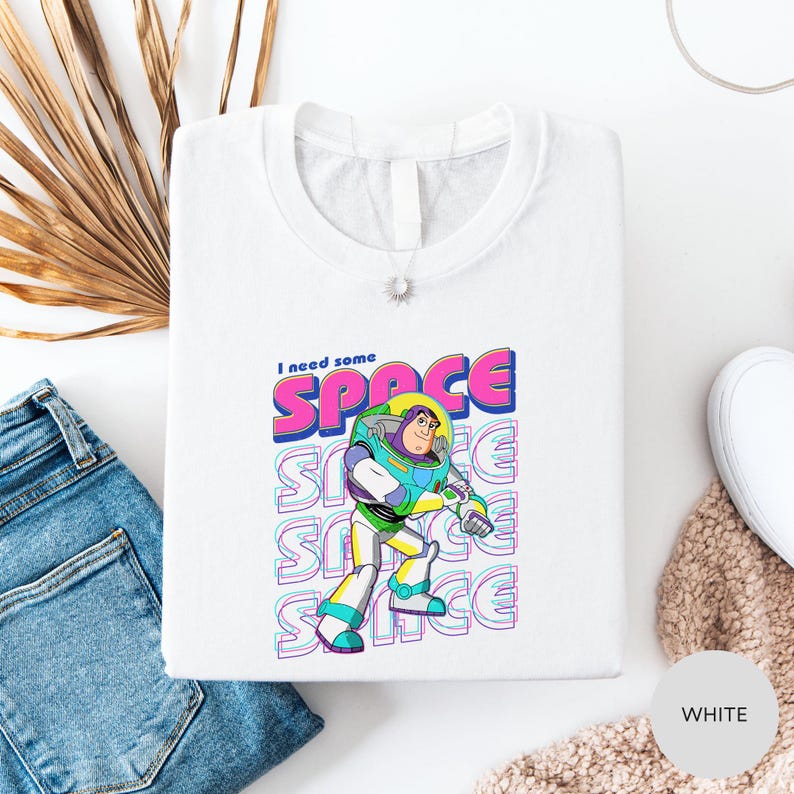 a white t-shirt with a colorful graphic of a character from the Toy Story franchise, along with the text "I need some SPACE" printed on it. The t-shirt is laid out on a white surface, with a pair of blue jeans and a beige sweater visible in the background.