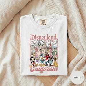 A white t-shirt with a graphic design of Disney characters and landmarks, including Mickey Mouse, Minnie Mouse, Donald Duck, and Goofy, along with the text "Disneyland Est. 1955" and "California".
