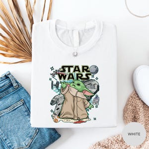 a white t-shirt with a Star Wars graphic design, including a depiction of the character Baby Yoda. The t-shirt is laid out on a white surface, along with a pair of blue jeans and a beige sweater.