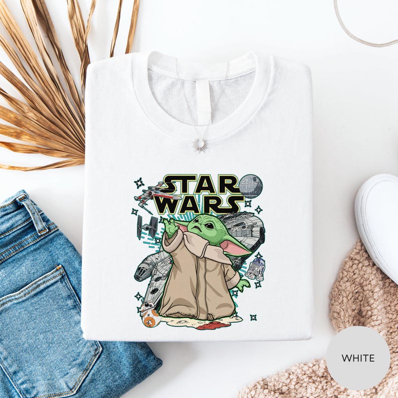 a white t-shirt with a Star Wars graphic design, including a depiction of the character Baby Yoda. The t-shirt is laid out on a white surface, along with a pair of blue jeans and a beige sweater.