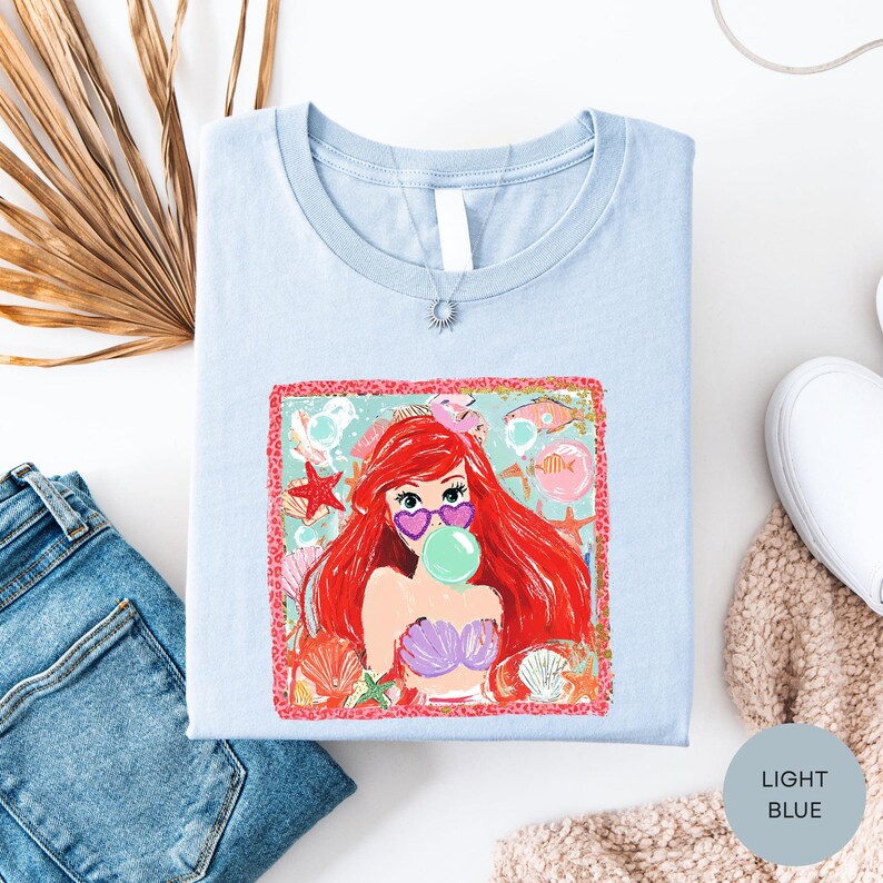 a light blue t-shirt with a colorful graphic of a mermaid, a pink and purple flower, and a starfish. The t-shirt is laid out on a white surface, along with a pair of blue jeans and a white sweater.
