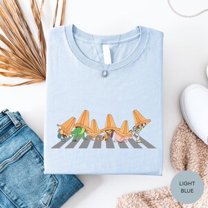 a light blue t-shirt with a graphic of cartoon characters wearing sombreros, which are orange cone-shaped hats, crossing a crosswalk. The t-shirt is laid out on a white surface, along with a pair of blue jeans and a white sweater.