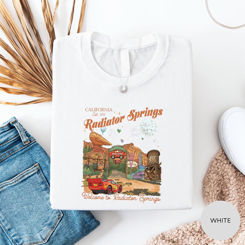 a white t-shirt with a graphic design depicting a desert scene with a red car, a wagon, and a cactus, along with the text "California Est. 2011 Radiator Springs Welcome to Radiator Springs".