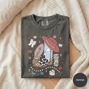A gray t-shirt with a colorful and whimsical design featuring a cowboy hat, flowers, stars, and other playful elements.