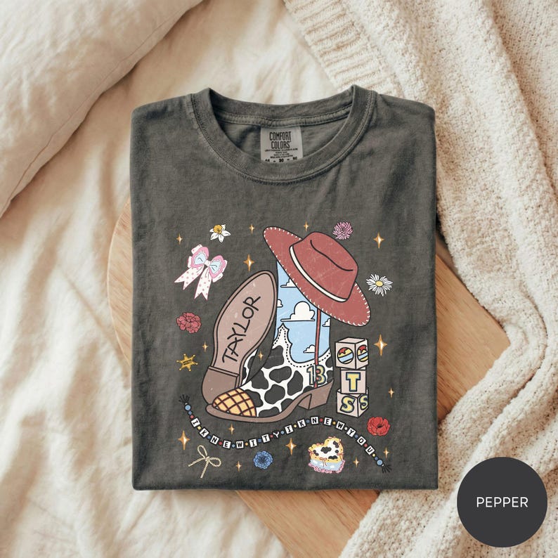 A gray t-shirt with a colorful and whimsical design featuring a cowboy hat, flowers, stars, and other playful elements.