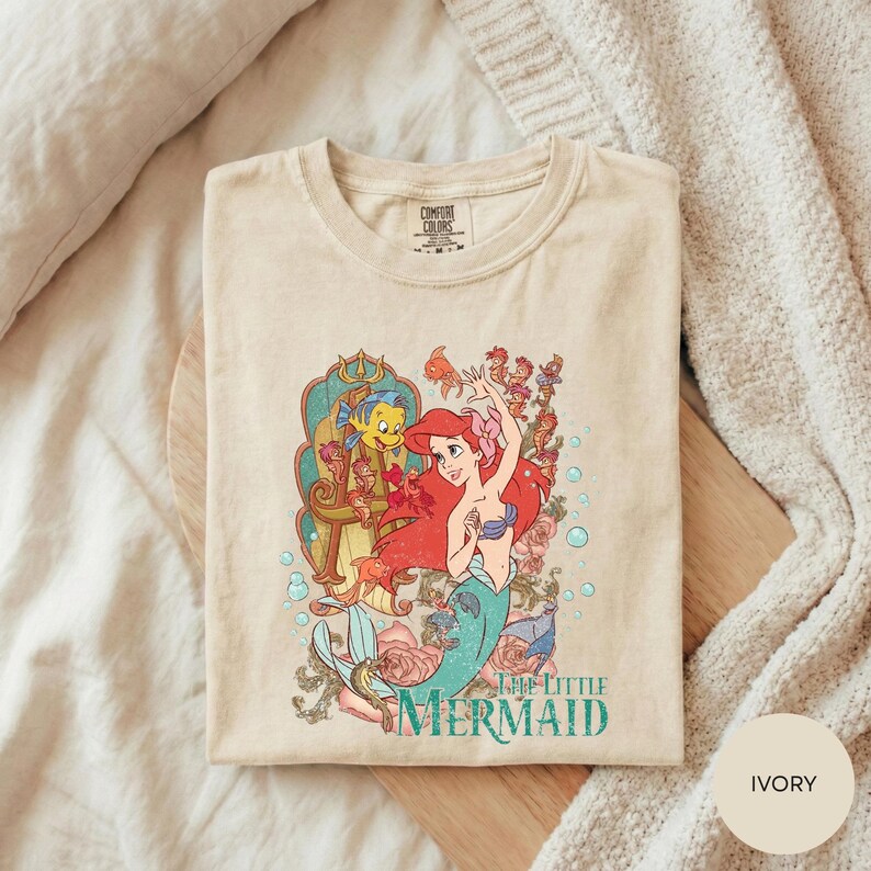 A t-shirt featuring the character Ariel from the Disney film "The Little Mermaid" is displayed on a wooden surface, with a beige blanket in the background.