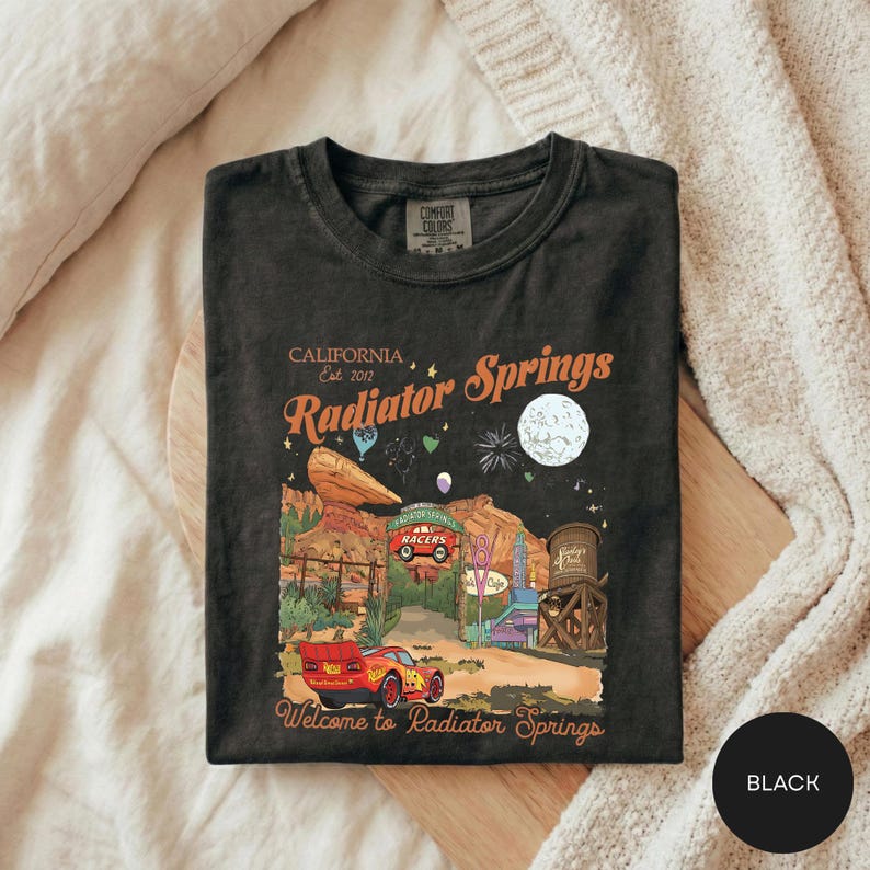 A black t-shirt with a graphic design depicting a desert landscape, a red car, and various other elements associated with the fictional world of the movie "Cars".