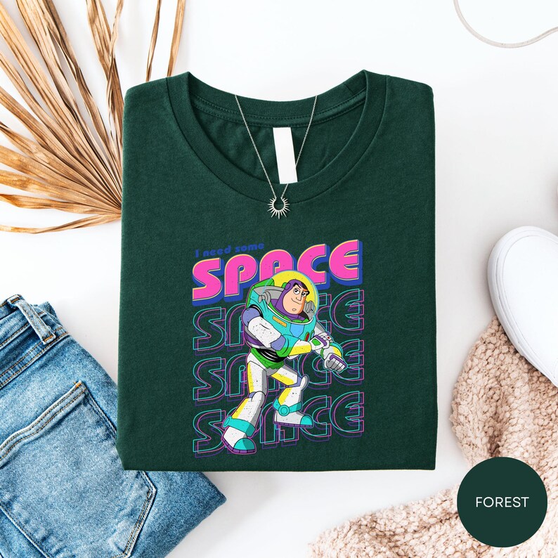 a green t-shirt with a graphic of a character from the Toy Story franchise, specifically Buzz Lightyear, along with the text "I need some space" printed on it. The t-shirt is laid out on a white surface, accompanied by a pair of jeans, a necklace, and a beige sweater.