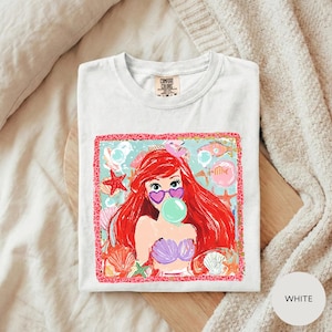 A white t-shirt with a colorful graphic design featuring a young girl with red hair blowing a bubble gum bubble.