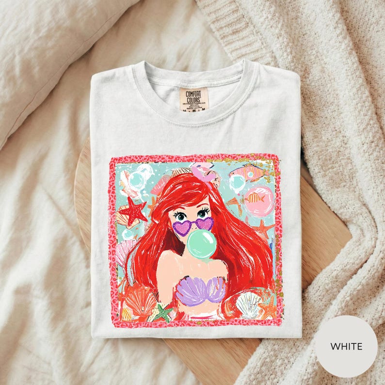 A white t-shirt with a colorful graphic design featuring a young girl with red hair blowing a bubble gum bubble.