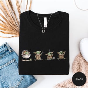 a black t-shirt with a graphic design of four baby alien characters, likely representing the characters from the Star Wars franchise. The t-shirt is laid out on a white surface, along with a pair of blue jeans and a beige sweater.