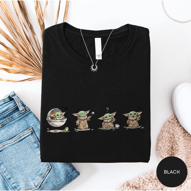 a black t-shirt with a graphic design of four baby alien characters, likely representing the characters from the Star Wars franchise. The t-shirt is laid out on a white surface, along with a pair of blue jeans and a beige sweater.