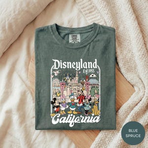 A green t-shirt with a graphic design of Disney characters and the text "Disneyland Est. 1955" printed on it, laid out on a wooden surface.