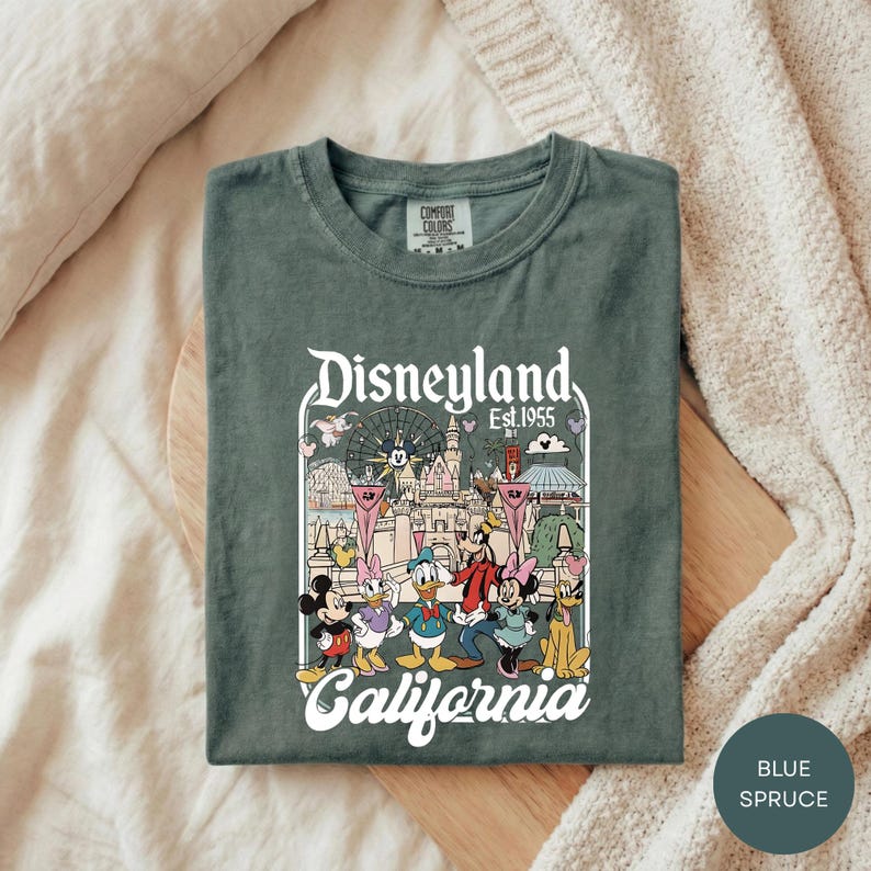 A green t-shirt with a graphic design of Disney characters and the text "Disneyland Est. 1955" printed on it, laid out on a wooden surface.