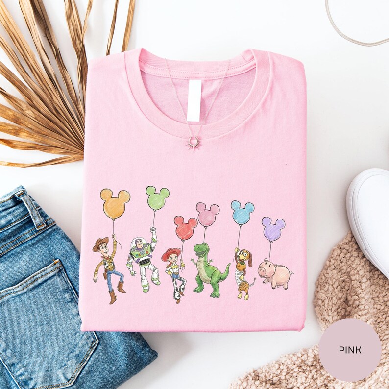 a pink t-shirt with a cartoon character design, including a character holding a balloon, a character holding a balloon, and a character holding a balloon. The t-shirt is placed on a white surface, along with a pair of jeans and a beige sweater.