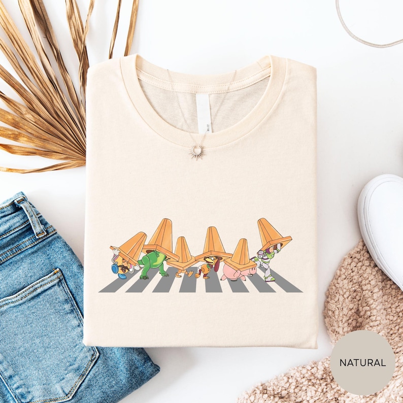 a white t-shirt with a graphic design of cartoon characters wearing orange hats, standing on a crosswalk. The t-shirt is placed on a white surface, along with a pair of blue jeans and a beige sweater.
