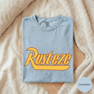A light blue t-shirt with a yellow and orange graphic design featuring the word "Rusteze" printed on it, laid out on a wooden surface.