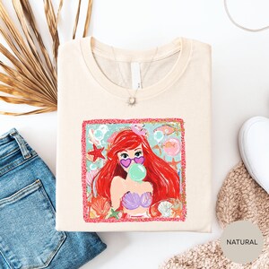 a white t-shirt with a colorful graphic of a mermaid character, along with a pair of blue jeans and a beige sweater.