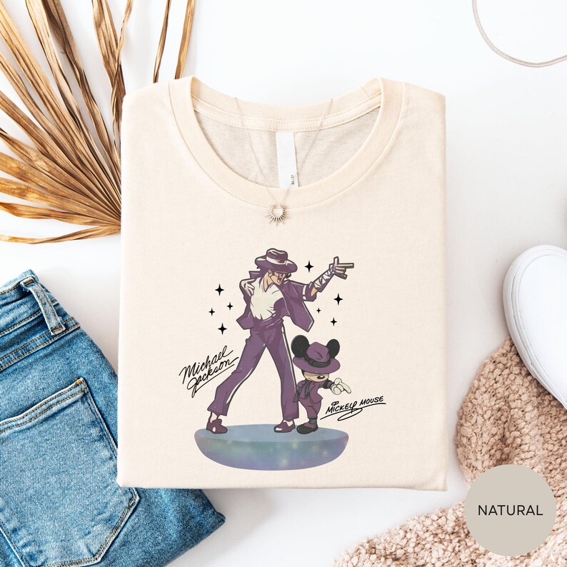 a white t-shirt with a graphic design of a man and a child, along with the text "Michael Jackson" and "Mackie Mouse". The t-shirt is laid out on a white surface, with a pair of blue jeans and a beige sweater visible in the background.