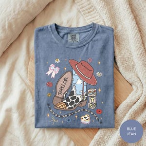 A blue t-shirt with a cartoon-style illustration of a cowboy hat, a bow, and various other objects, along with the word "Taylor" written on it. The t-shirt is laid out on a bed, and there is a blanket visible in the background.