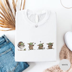 a white t-shirt with a graphic design depicting three baby alien characters, likely representing the characters from the Star Wars franchise. The t-shirt is laid out on a white surface, with a pair of blue jeans and a white sweater visible in the background.