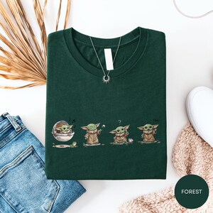 a green t-shirt with a graphic design of four baby alien characters, each with a unique pose, accompanied by a necklace and a pair of jeans.