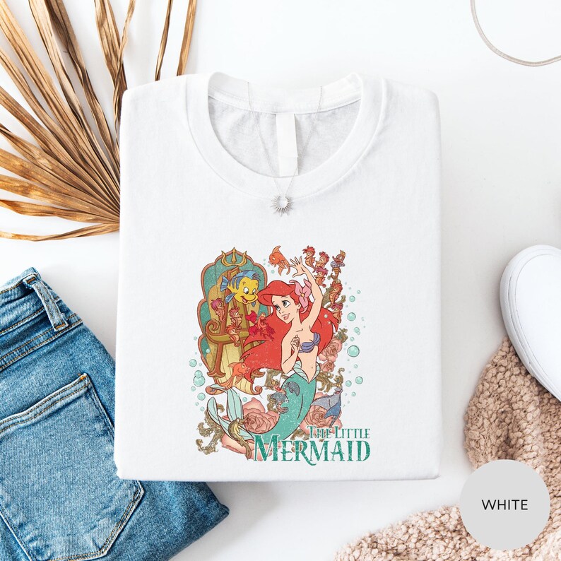 a white t-shirt with a colorful graphic of the mermaid character from the Disney animated film "The Little Mermaid". The t-shirt is laid out on a white surface, along with a pair of blue jeans and a beige knitted item.