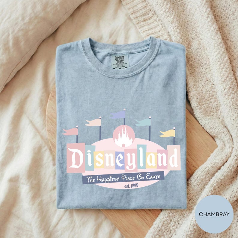 A blue t-shirt with a Disneyland logo and text, laid out on a wooden surface with a beige blanket.