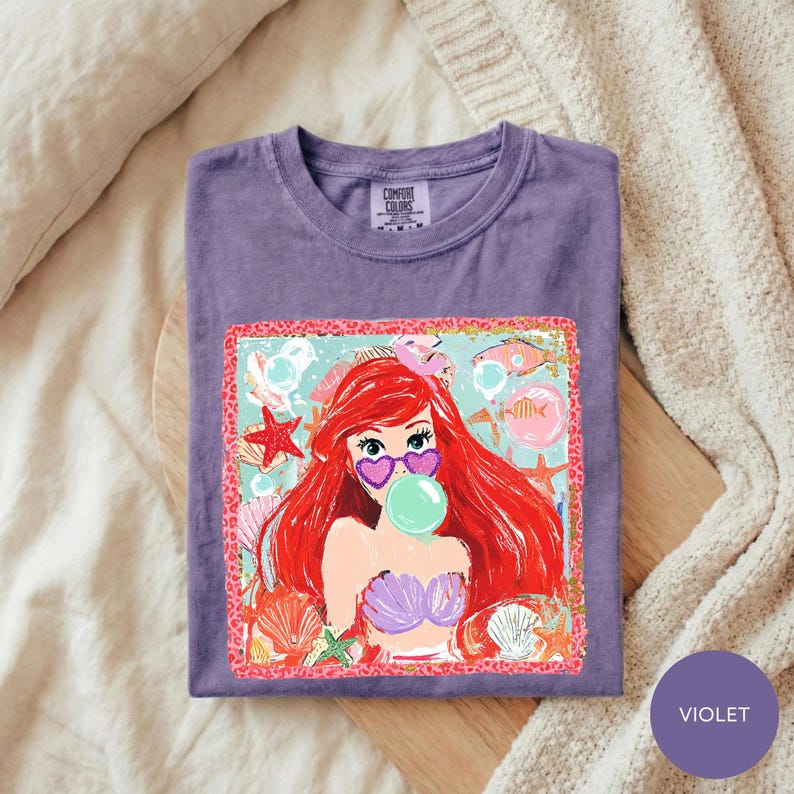 A purple t-shirt with a colorful graphic of a mermaid character blowing a bubble gum bubble, surrounded by seashells and starfish.