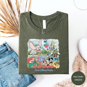 A green t-shirt with a Disney-themed graphic, including characters from the Disney film "Magic Kingdom" and a castle, is displayed alongside a pair of jeans and a sweater.
