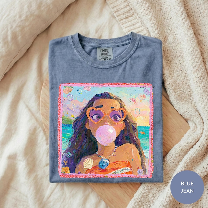 A blue t-shirt with a colorful illustration of a young girl wearing sunglasses and blowing a bubble gum bubble.
