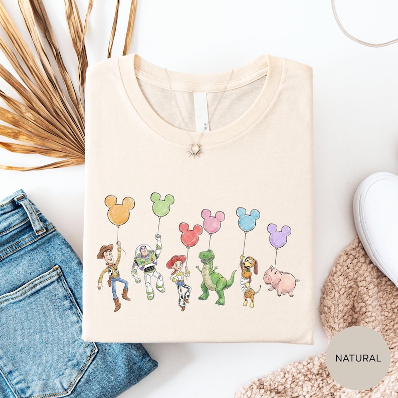 a t-shirt with a cartoon character design, including a character holding a balloon, a character holding a balloon, and a character holding a balloon. The t-shirt is placed on a white surface, along with a pair of jeans and a beige sweater.