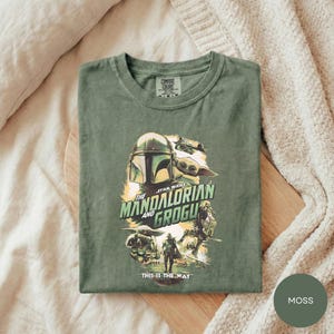 A green t-shirt with a graphic design featuring characters from the Star Wars series, specifically The Mandalorian and the Frogs.