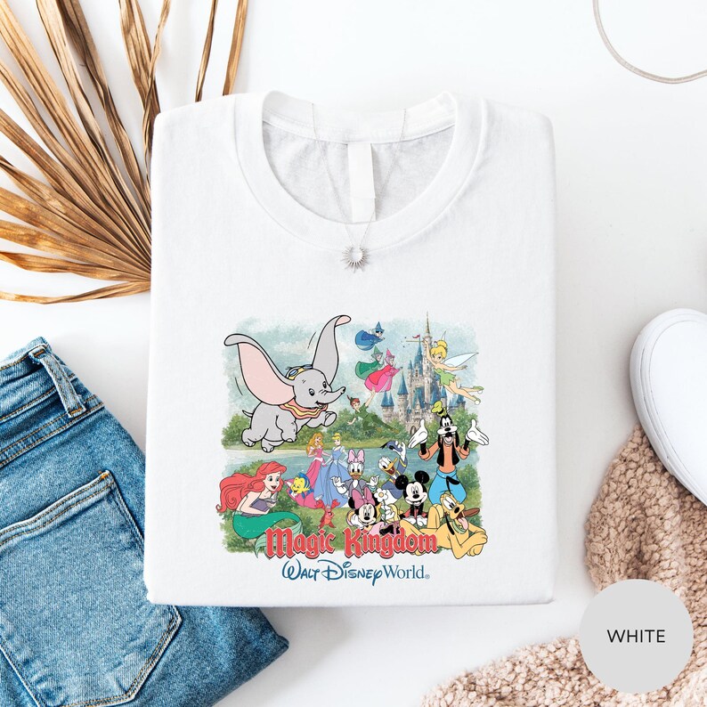 A white t-shirt with a colorful graphic of Disney characters from the Magic Kingdom, including Dumbo, Minnie Mouse, Mickey Mouse, Donald Duck, and other iconic figures, along with a castle and palm trees.