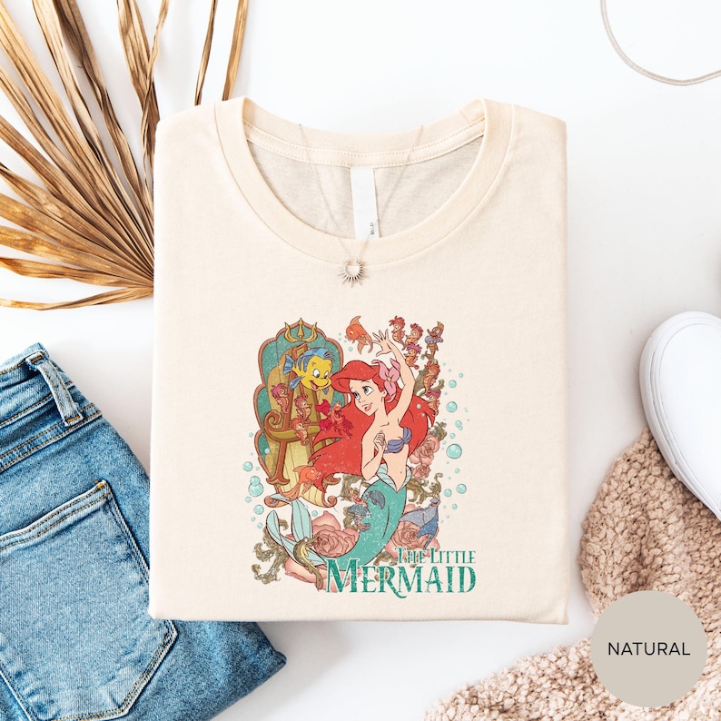 a white t-shirt with a colorful graphic of the mermaid character from the Disney film "The Little Mermaid". The t-shirt is laid out on a white surface, along with a pair of blue jeans and a beige sweater.