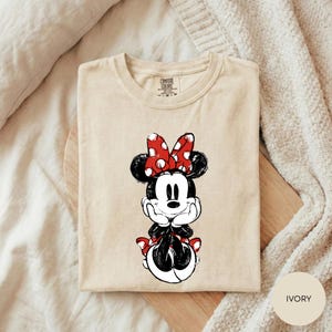 A beige t-shirt with a Minnie Mouse graphic printed on it, placed on a wooden surface.