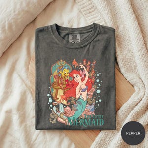 A gray t-shirt with a colorful graphic of the mermaid character Ariel from the Disney film "The Little Mermaid" printed on it, along with the text "The Little Mermaid".