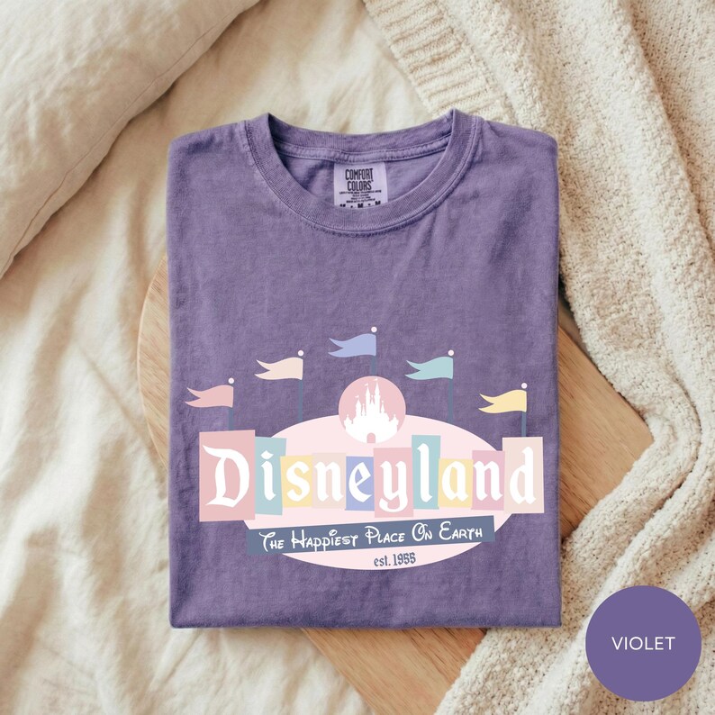 A purple t-shirt with a Disneyland logo and text, laid out on a bed with a white blanket.