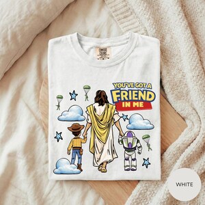A white t-shirt with a graphic design depicting a person in a yellow robe and a child in a cowboy hat, along with a character in a purple suit and a character in a blue suit, all holding hands. The text on the t-shirt reads "You've got a FRIEND in ME".