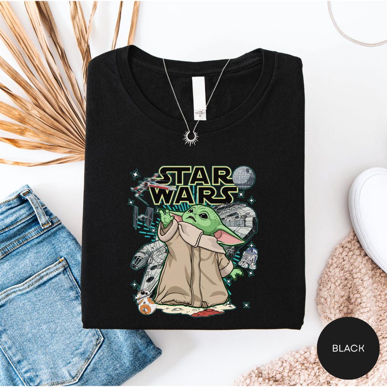 a black t-shirt with a Star Wars graphic design, including a depiction of the character Baby Yoda. The t-shirt is laid out on a white surface, along with a pair of blue jeans and a beige sweater.