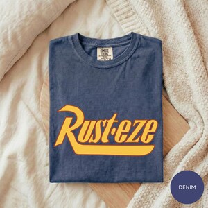 A blue t-shirt with the word "Rusteze" printed in yellow text is displayed on a wooden surface, with a white blanket and a denim-looking item in the background.