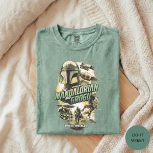 A green t-shirt with a graphic design featuring characters from the Star Wars series, specifically The Mandalorian and the Frogs.