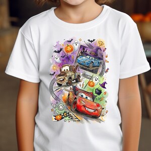 May include: A white short-sleeve t-shirt featuring a colorful graphic print of animated race cars, including a red car, a blue car, and a brown tow truck, surrounded by Halloween elements like ghosts, bats, pumpkins, and purple witch hats.