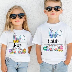 May include: Two children in white t-shirts with Easter designs. The shirts display bunny ears, names, Easter eggs, and bunny feet. The girl's shirt says "Jessie" and the boy's shirt says "Caleb". Both children are wearing sunglasses.