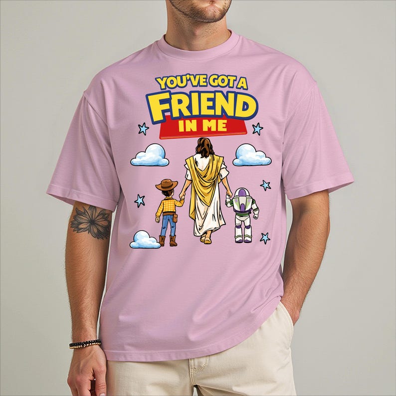A man wearing a pink t-shirt with a graphic design featuring a cartoon depiction of Jesus Christ, a woman, and a child. The text on the t-shirt reads "You've Got a Friend in Me".
