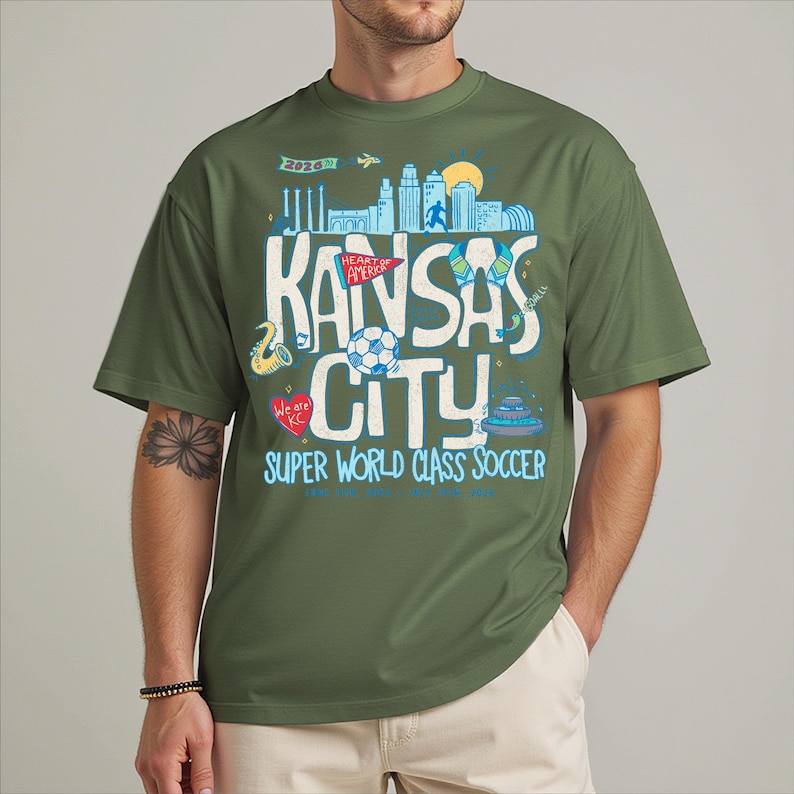 A man wearing a green t-shirt with a graphic design featuring various elements related to Kansas City, including a city skyline, a soccer ball, and a heart symbol.