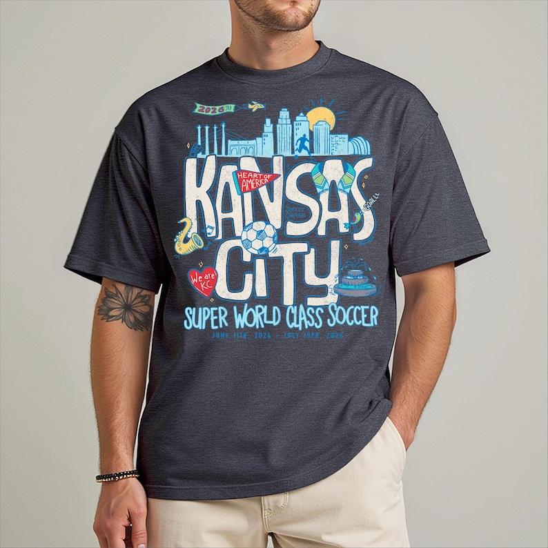 A man wearing a gray t-shirt with a graphic design featuring various elements related to Kansas City, including a city skyline, a soccer ball, and other sports-related imagery.